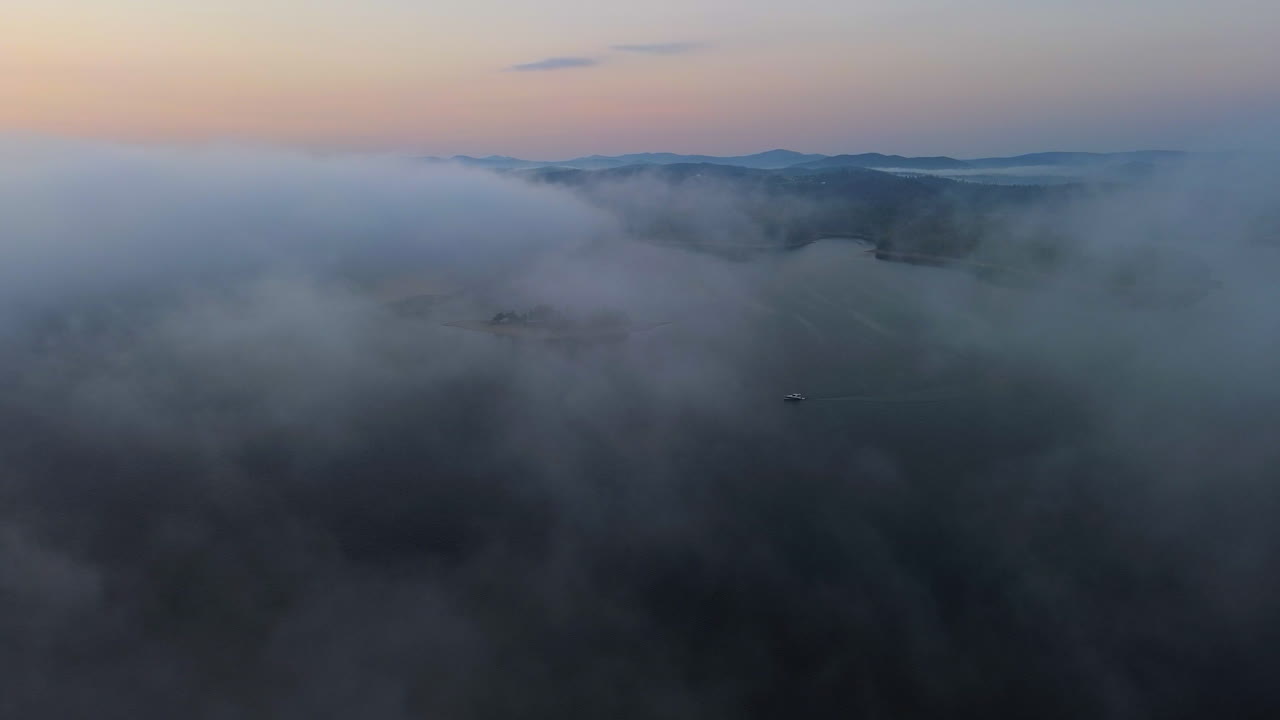 Sailboat lost in thick fog drifting over mountain lake at sunrise - dreamlike aerial wide shot. Gentle light illuminates distant hills. Solina, Poland