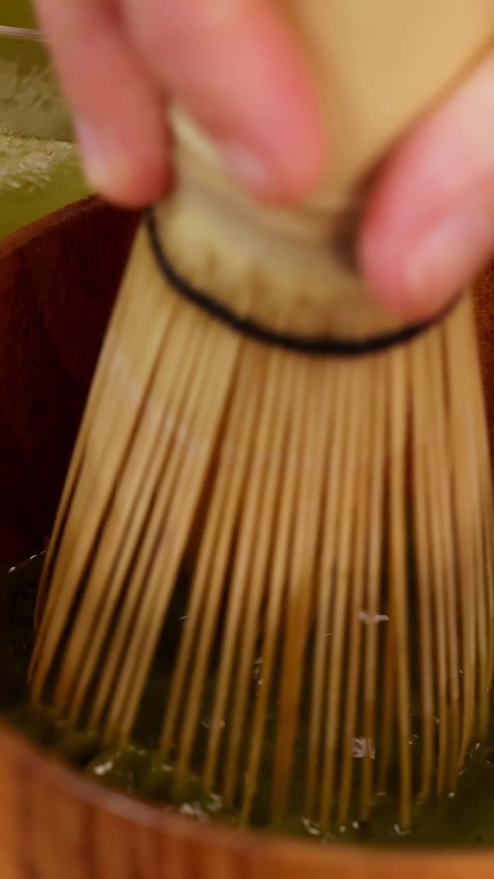 Primo piano di una mano che sbatte un vivace tè matcha in una ciotola di legno sotto una morbida illuminazione da studio.