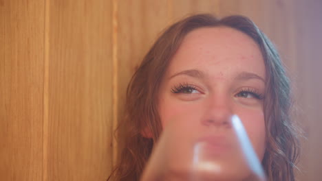 close up of young woman toasting glass with friend while sipping red juice in wood paneled interior, soft backlight casting warm haze over wet curls and serene smile