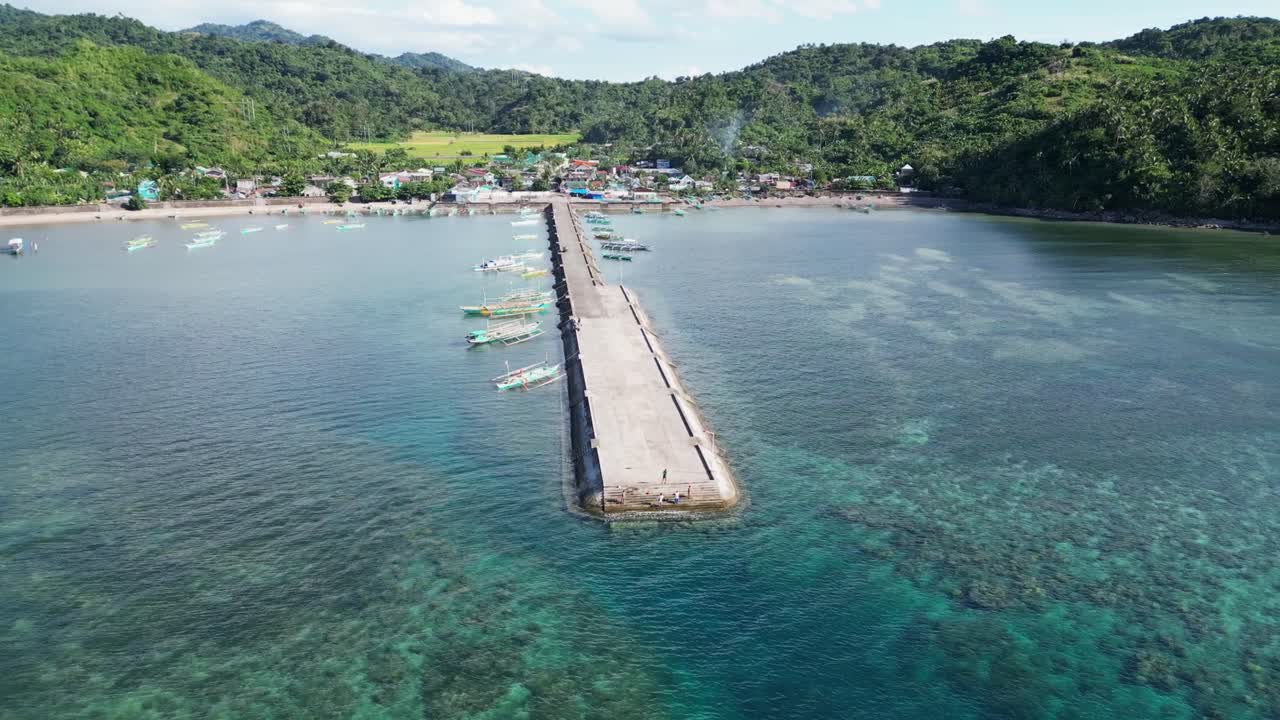 시네마틱 에어리얼 풀백  ⁇ 도 뷰 블루 아웃리거  ⁇ 카 보트가 열대 마을 도크에 도착하여 조용한 해변 마을과 울창한 산을 배경으로합니다.