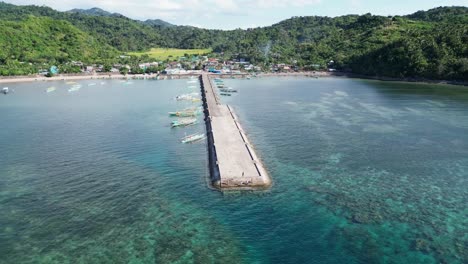 cinematic aerial pullback orbit view of blue outrigger bangka boat arriving at tropical village dock with serene seaside town and lush mountains in background