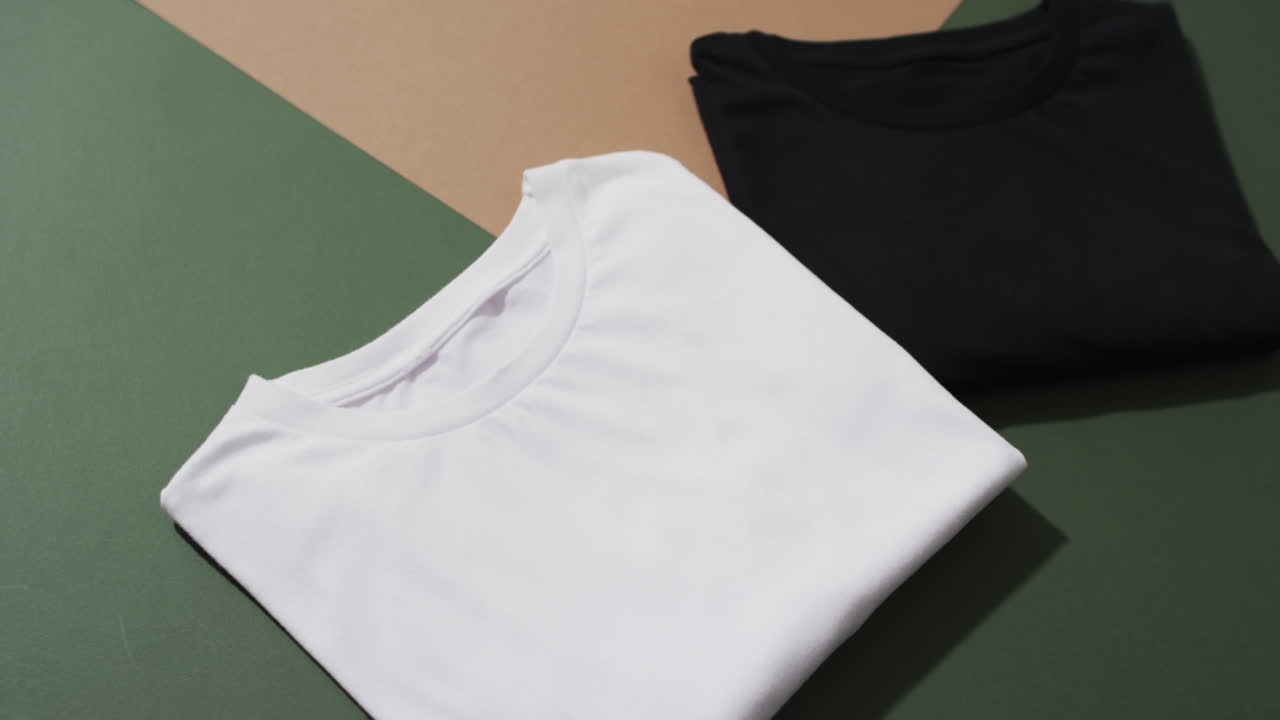Video of flat lay of folded white and black t shirts with copy space on green and brown background