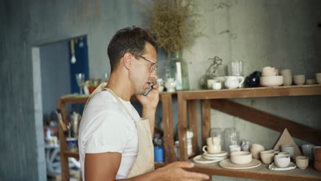 a happy young potter in glasses is talking on a smartphone while standing near his clay works. traditional business concept