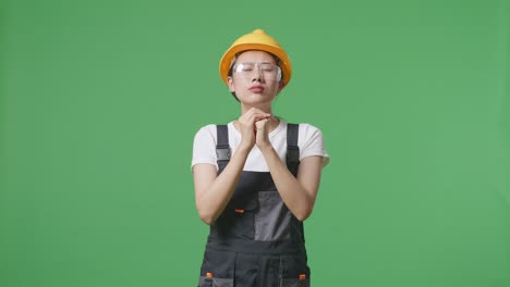 asian woman worker wearing goggles and safety helmet praying for something while standing in the green screen background studio