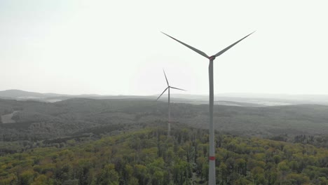 Aerial-dolly-shot-of-a-couple-of-wind-turbines-surrounded-by-forest,-bright-daylight