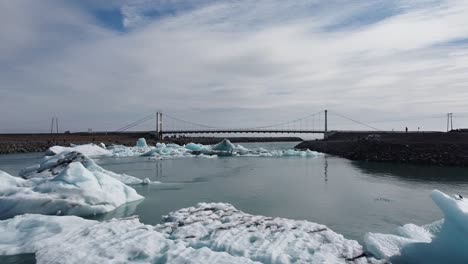 Jokulsarlon-Glacier-Lagoon-with-icebergs-in-Iceland,-aerial-view-above-melting-ice-towards-suspension-bridge