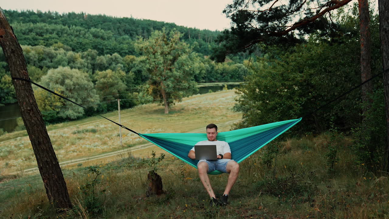 Man working relaxed in the blue hammock hanging among the trees. Freelancer working remotely on his laptop.