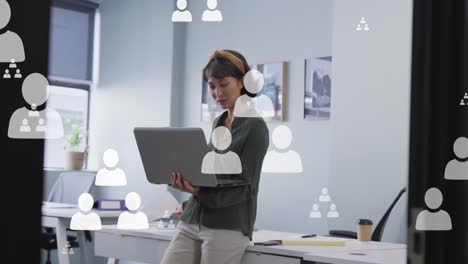 Animation-of-multiple-profile-icons-floating-against-asian-woman-using-laptop-at-office