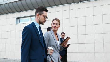 Caucasian-young-businesswoman-and-businessman-walking-down-street-while-talking-and-using-a-tablet-and-holding-coffee-to-go