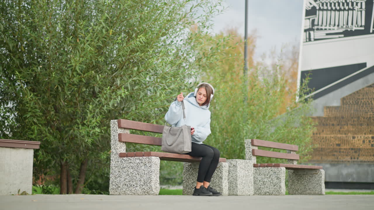 Fair lady walking with handbag, sitting down on concrete bench in outdoor park setting, background includes lush green leaves, concrete benches, and modern building, relaxed urban environment