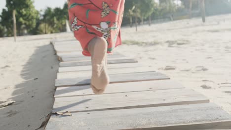 low section of biracial woman in red dress walking barefoot on boardwalk on sunny beach, slow motion