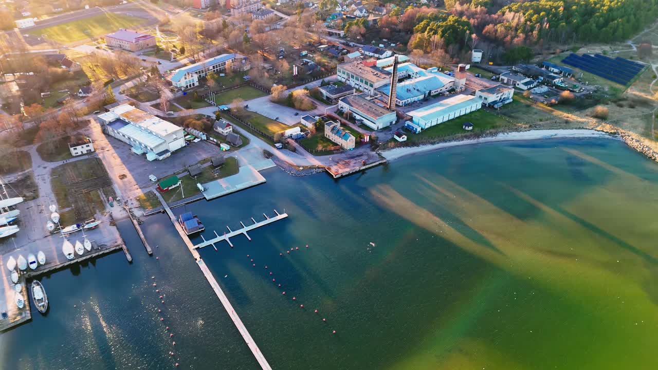 Autumnal aerial view of Engure Port, Latvia, bathed in warm, golden hour sunlight, highlighting the varying blue-green hues of the water, a bustling marina, and industrial buildings along the shore