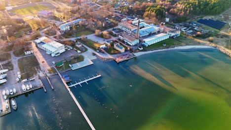 autumnal aerial view of engure port, latvia, bathed in warm, golden hour sunlight, highlighting the varying blue-green hues of the water, a bustling marina, and industrial buildings along the shore