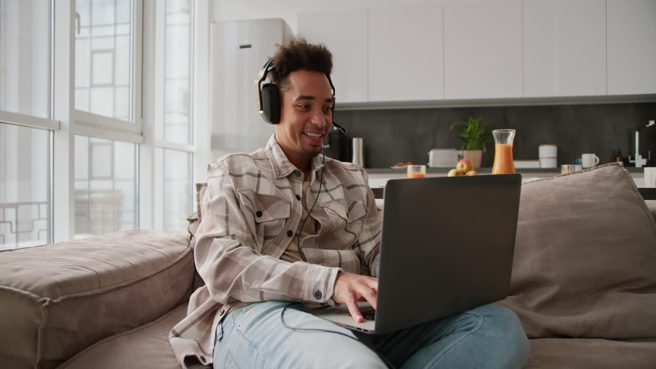 A happy Black skinned young brunette man with stubble wearing black modern headphones and a cream plaid shirt plays a video game on his gray laptop while sitting on a modern sofa in a modern studio apartment