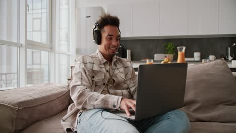 A-happy-Black-skinned-young-brunette-man-with-stubble-wearing-black-modern-headphones-and-a-cream-plaid-shirt-plays-a-video-game-on-his-gray-laptop-while-sitting-on-a-modern-sofa-in-a-modern-studio-apartment