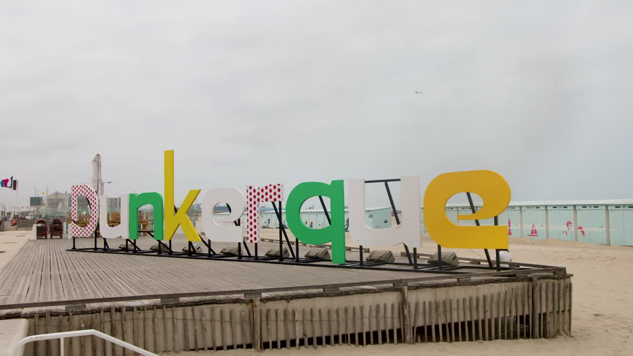 Static wide shot of Dunkerque sign on sandy boardwalk, overcast sky, muted daylight, coastal environment