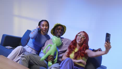 studio shot of group of gen z friends sitting on sofa posing for selfie with toy alien on mobile phone 2