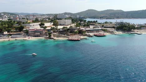 Drone-view-in-Albania-flying-sideways-in-front-of-white-sand-beaches,-blue-crystal-clear-water,-green-small-islands,-boats-and-hotels,-sunny-day-in-Ksamil