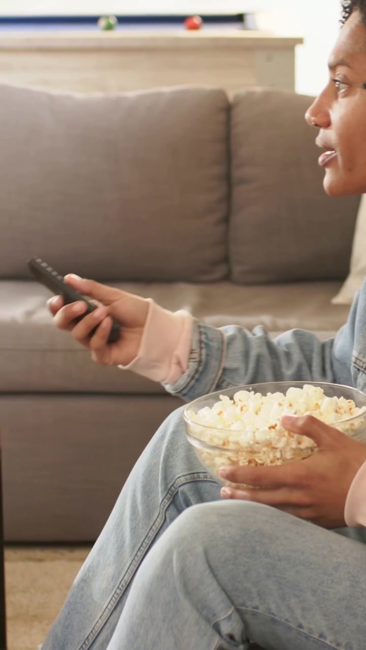 Video verticale: Uomo in denim che prende popcorn da una ciotola di vetro e tocca il telecomando in soggiorno, con spazio per il testo.