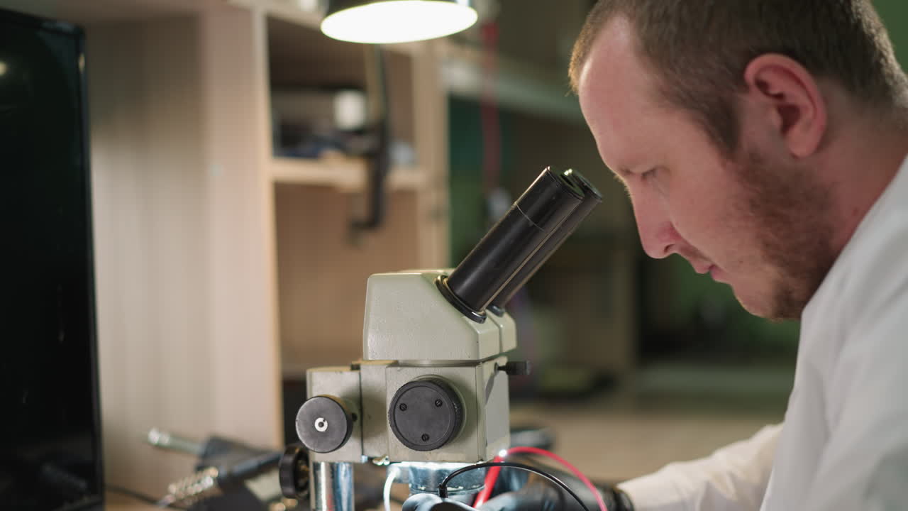 vista central de un técnico con una bata de laboratorio blanca y guantes negros probando cuidadosamente un circuito bajo un microscopio usando un voltímetro en su laboratorio, con la lámpara de escritorio encendida