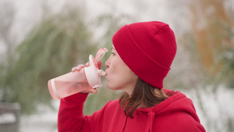 middle aged woman in red beanie and hoodie drinking water outdoors using pink bottle, set against blurred background with greenery and iron rail, while someone walks by in serene winter setting