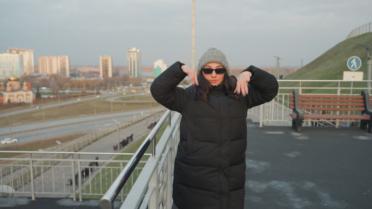 Donna sul balcone, balla e indica mentre fa vlogging, indossando piumino e cappello di lana, sistemandosi gli occhiali da sole. Coinvolgimento giocoso del pubblico con lo skyline urbano, energia invernale spontanea e gesti sicuri.