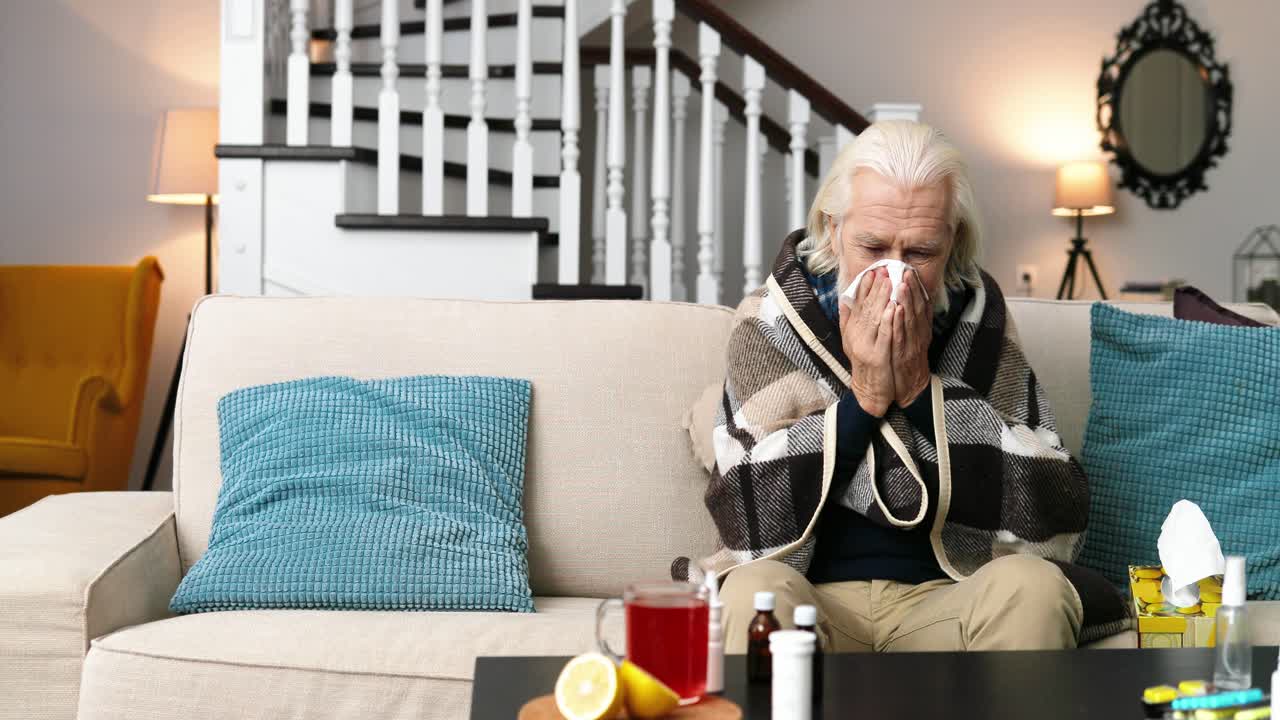 Senior man blowing his nose and sitting on the sofa