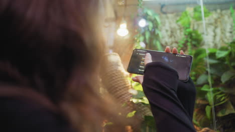 rear view of woman in black top adjusting phone settings while preparing to take photo of vibrant indoor greenery through glass enclosure, with warm lighting and lush leaves in background