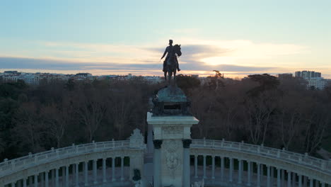 drone slowly flies backwards from alfonso xii statue at sunrise, revealing retiro gardens, lake, historic monument, park paths, city buildings, and beautiful morning cityscape panorama.