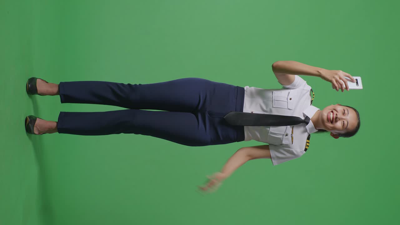 녹색 화면 배경 스튜디오에 서 있는 동안 스마트폰에서 비디오 통화를 하는 아시아 여성 조종사의 몸이 가득 차 있습니다.