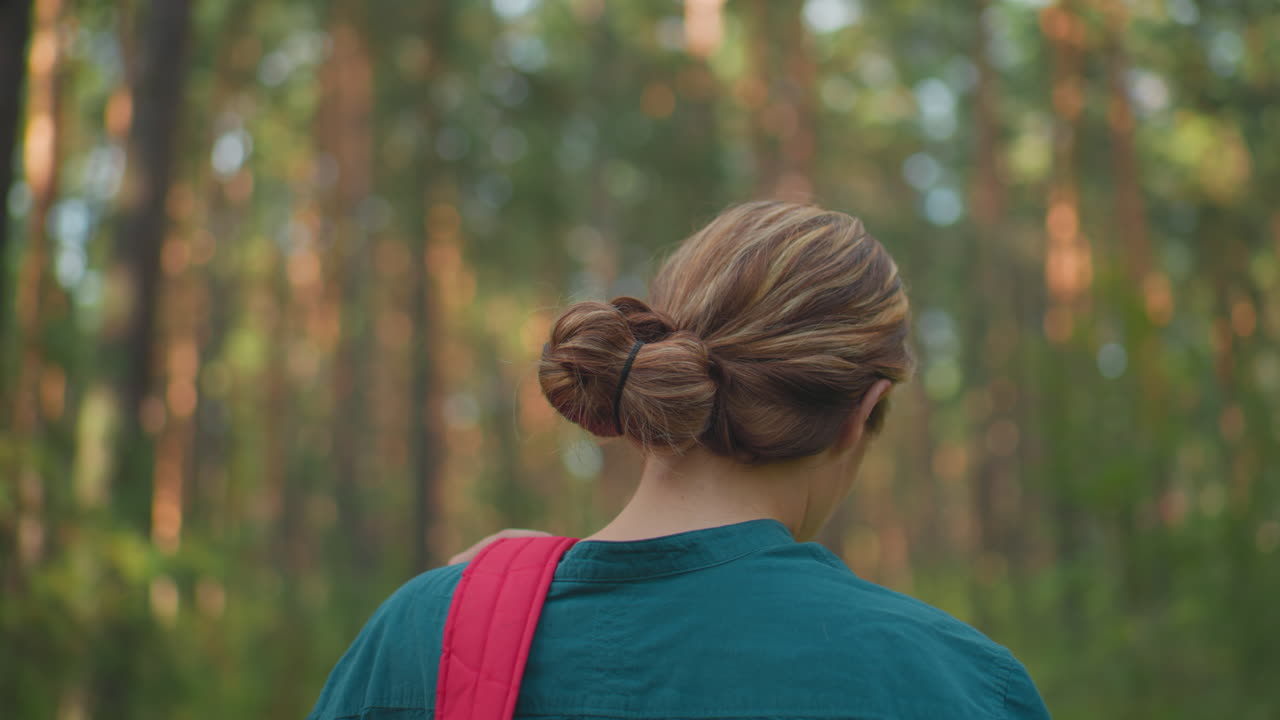 primer plano de una mujer caminando por un pacífico bosque iluminado por el sol con una mochila roja colgada sobre el hombro, ajustando la bolsa con la mano, su cabello está cuidadosamente atado hacia atrás, con el fondo ligeramente borroso