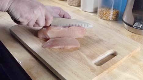 slow motion slider shot of slicing chicken breast pieces with a chef's knife on a wooden cutting board in a home kitchen