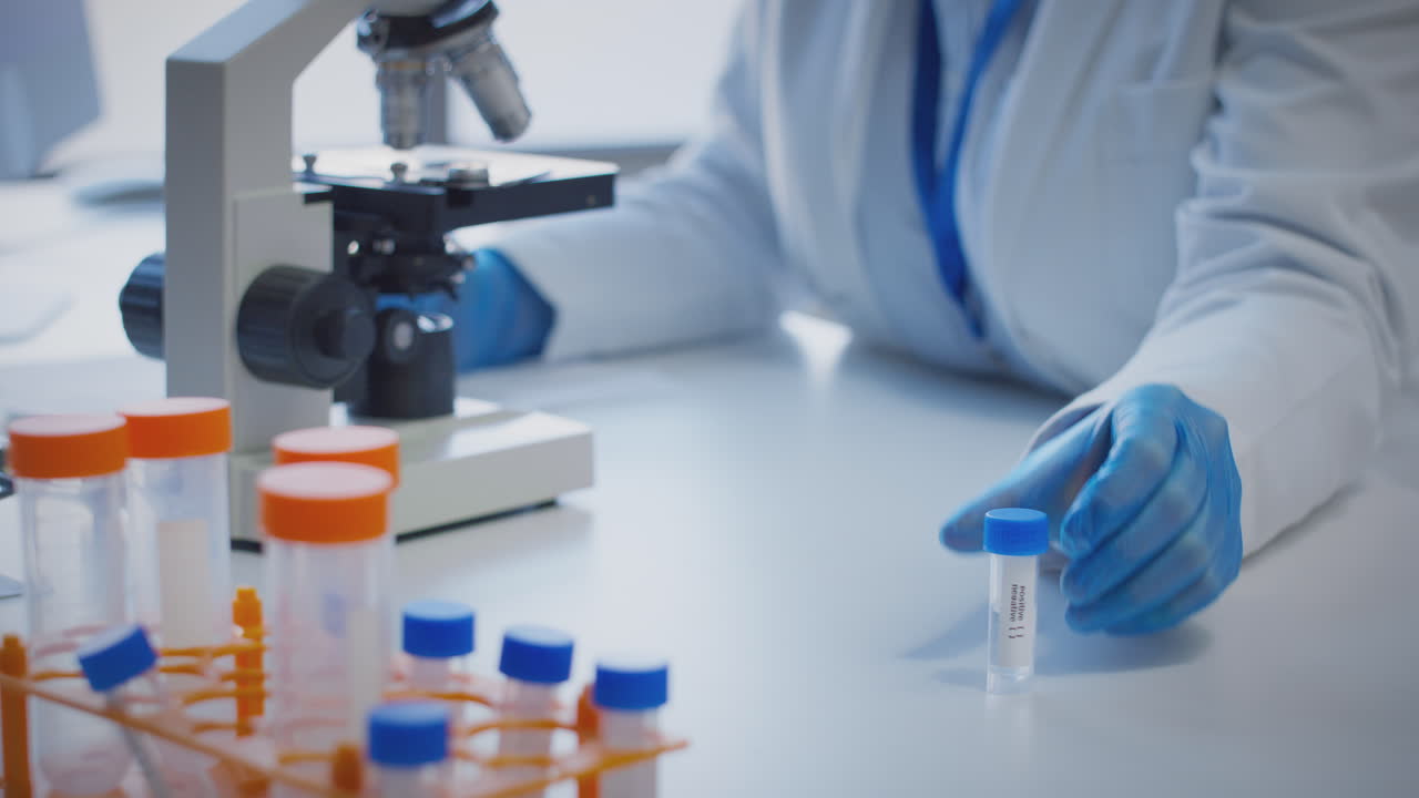 primer plano de un trabajador de laboratorio que lleva a cabo investigaciones utilizando un microscopio con un tubo de ensayo etiquetado como positivo y negativo