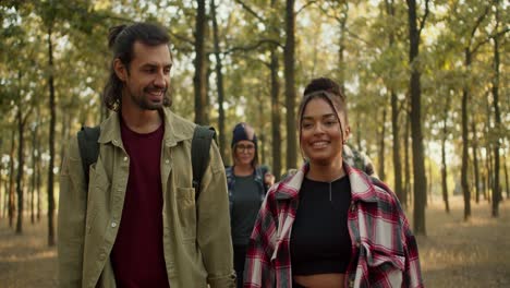 Happy-group-on-a-hike-front-view-man-and-girl-in-hiking-clothes-walking-along-the-summer-forest.-A-girl-with-Black-skin-in-a-checkered-shirt-walks-through-the-forest-and-communicates-with-a-brunette-man-with-stubble-in-a-light-green-jacket