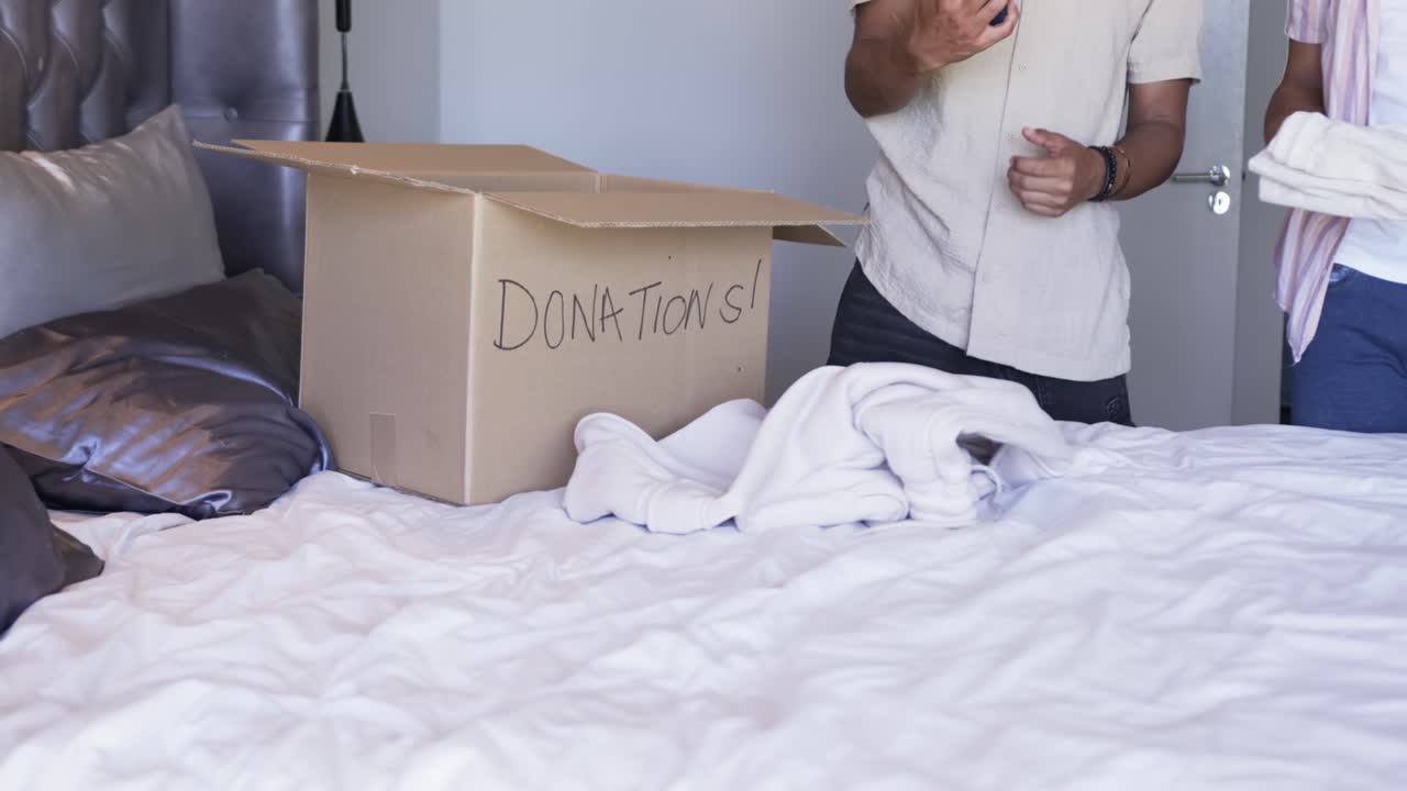 Divers mannelijk familieduo pakt telefoon op bed, controleert het scherm en pakt beddengoed in een donatiebox.
