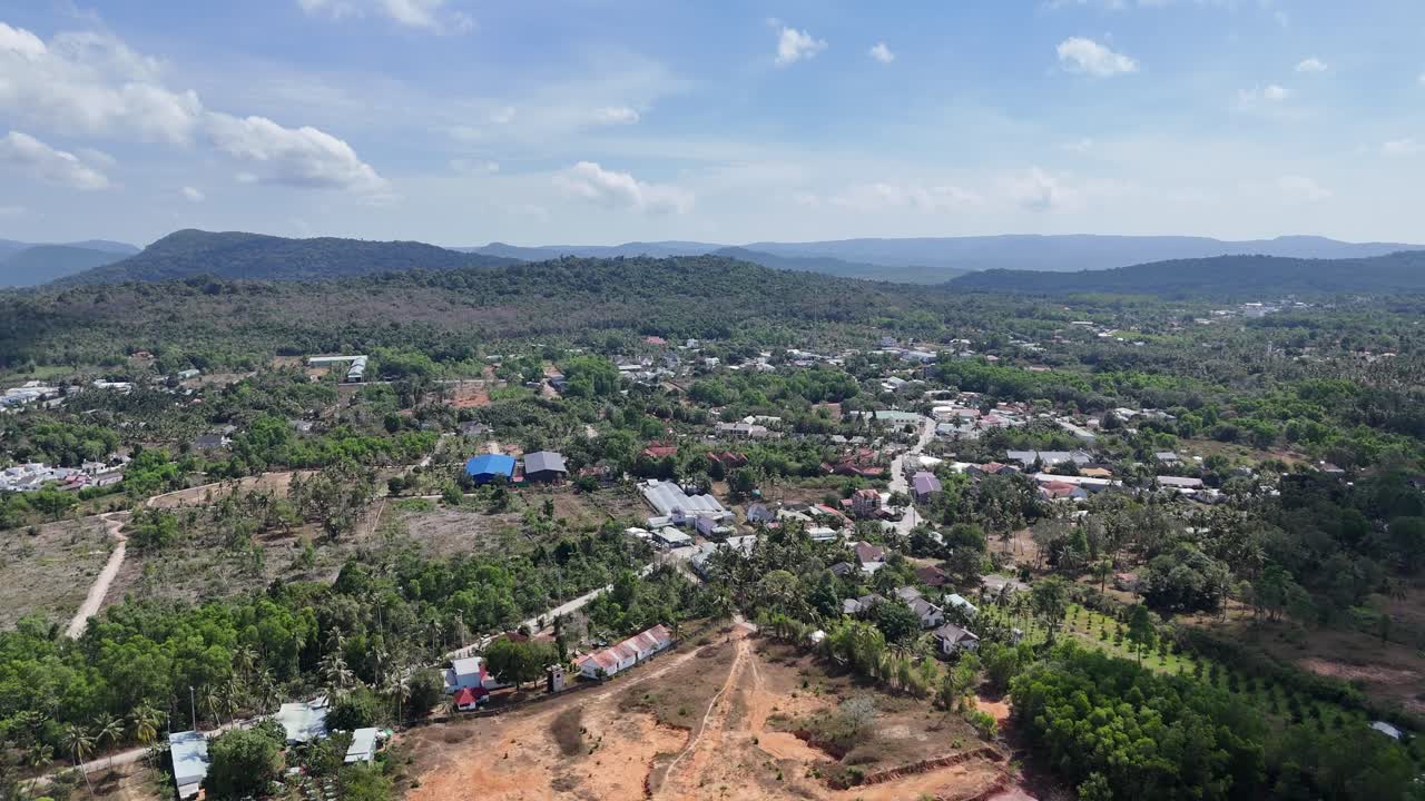 Aerial view of a road through dense vegetation with scattered residences and a distant body of water under a partly cloudy sky.