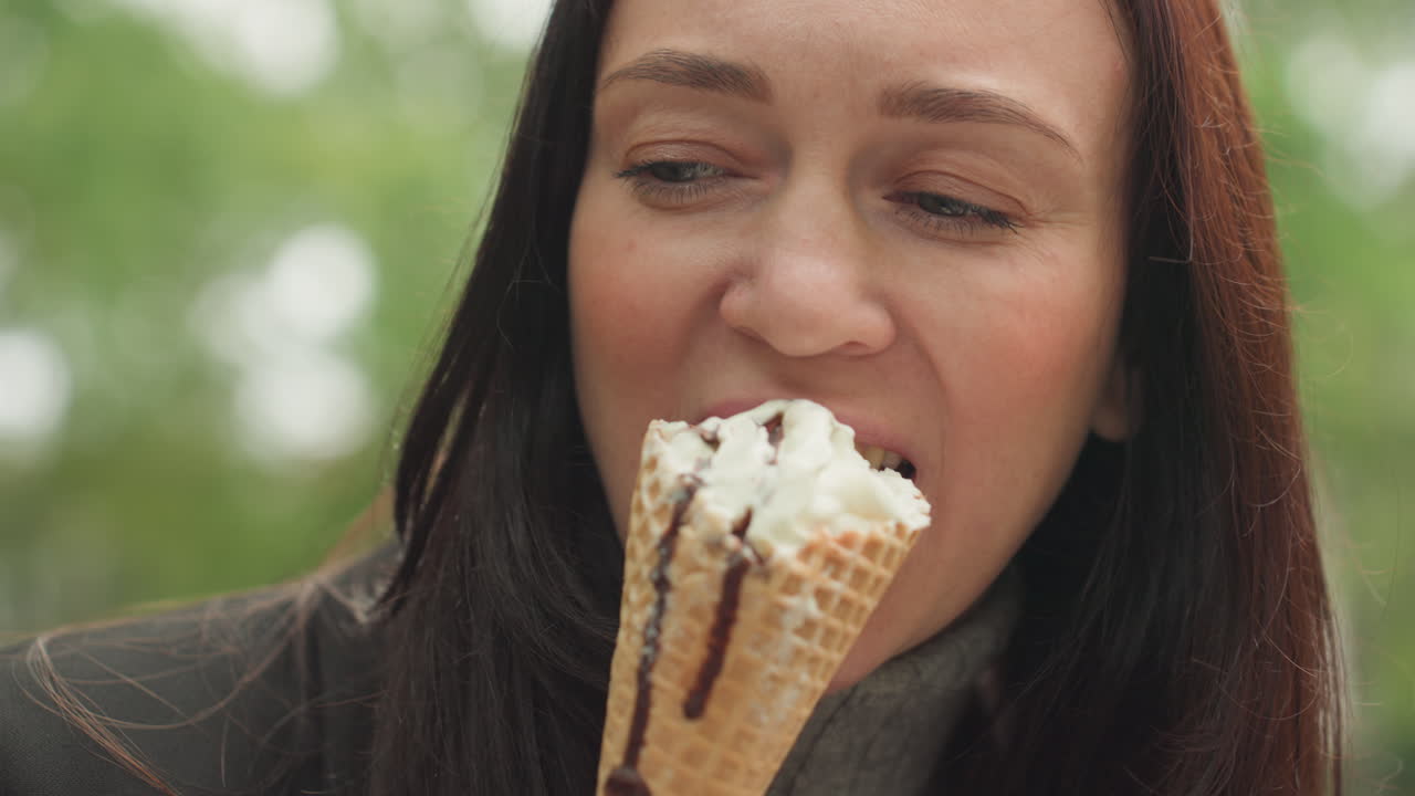 Woman eating ice cream cone closeup in park, waffle cone with vanilla and chocolate drizzle, contemplative expression, slow savoring bite, cool jacket and turtleneck, soft green foliage background