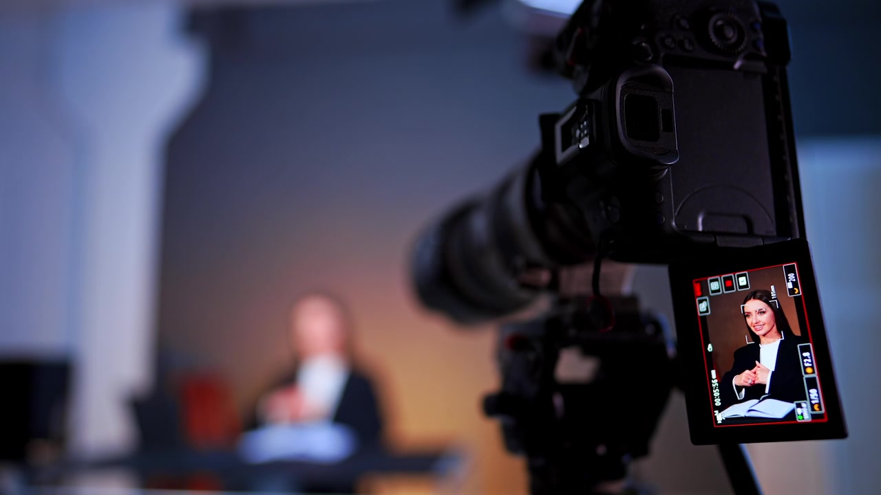 Telecamera con teleobiettivo che riprende una donna sorridente in un'intervista in studio. Attrezzatura con obiettivo lungo a fuoco con un'anteprima sullo schermo del soggetto e una figura sfocata sotto una luce calda.