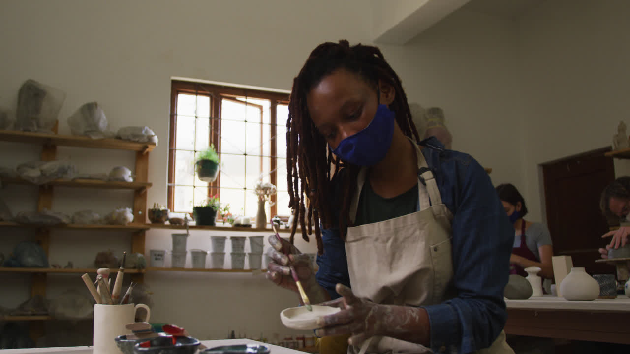 Female potter wearing face mask and apron using brush to paint on pottery at pottery studio
