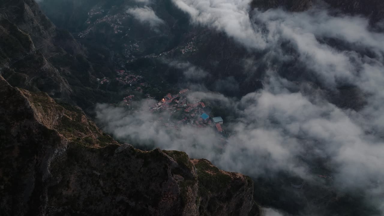 Drone shot of a small village nestled in a deep valley, partially covered by drifting clouds in Madeira’s mountains.