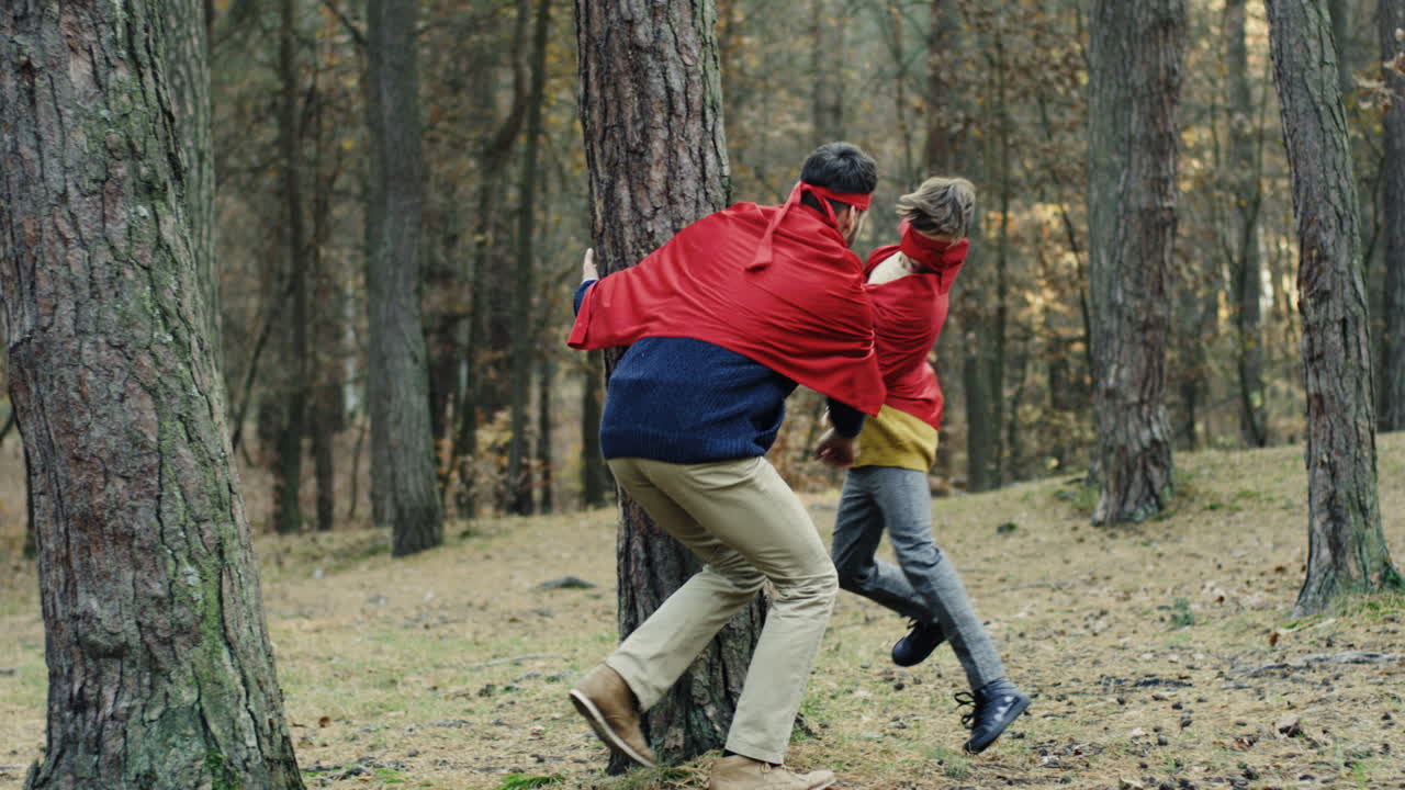 Caucasian cheerful father and son in red capes running in the forest and playing they being superheroes