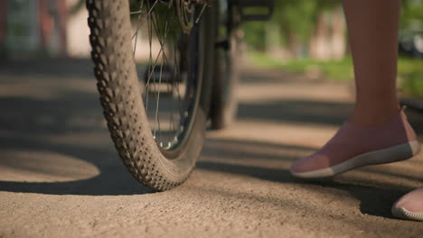 close-up of legs kicking a bicycle tire to check air pressure, the scene captures lush greenery and trees in the blurred background