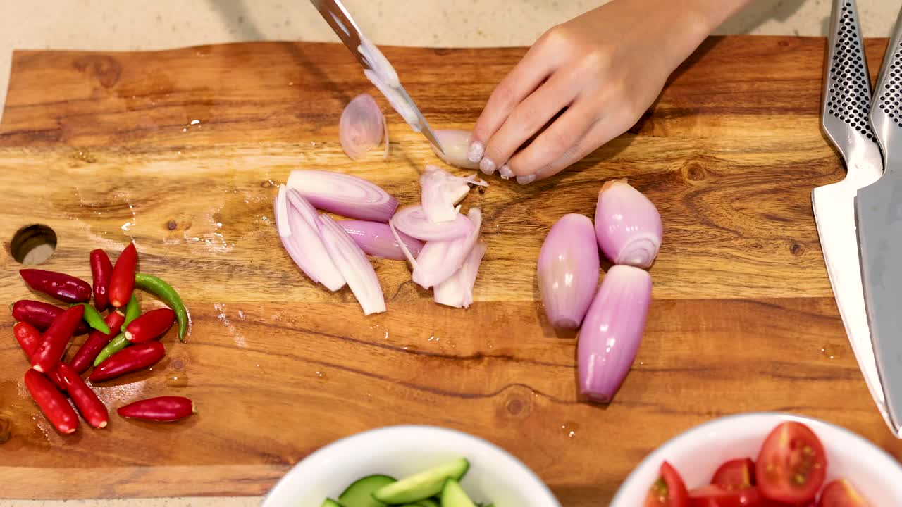 Hands slicing shallots on a wooden board with vibrant vegetables. Bright lighting, top-down view, emphasizing culinary preparation