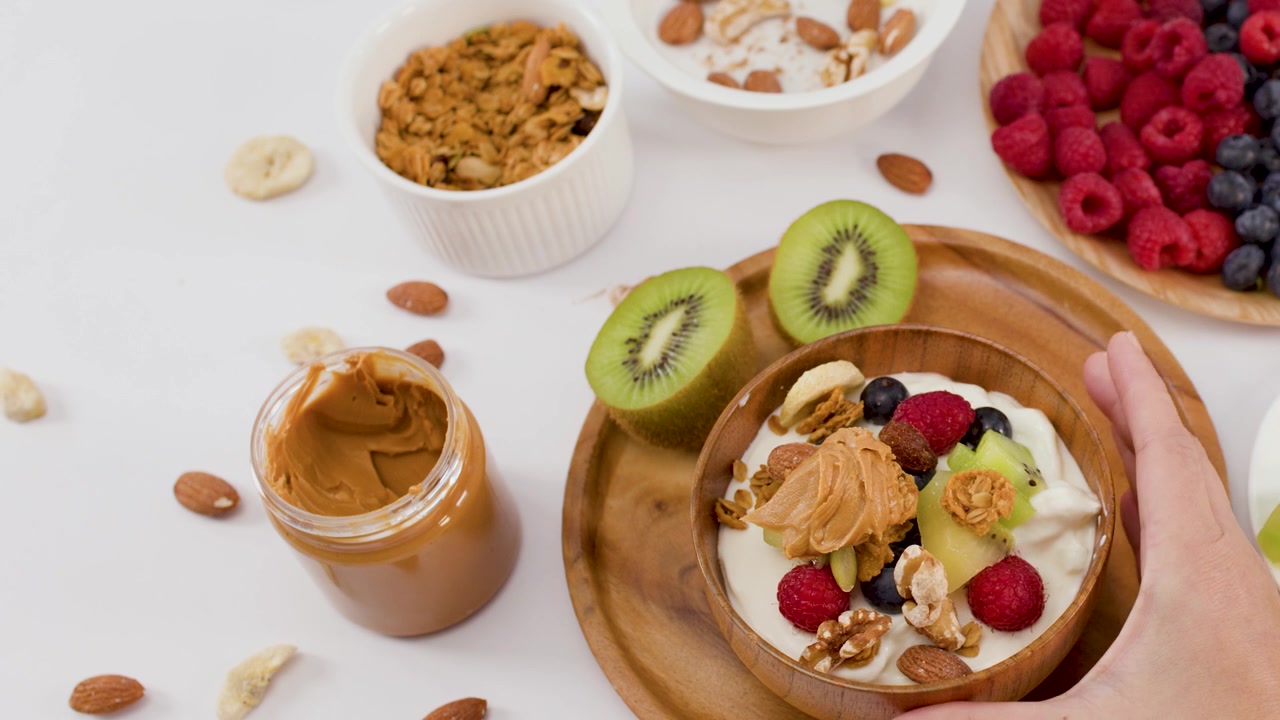 Top-down view of a hand lifting a wooden bowl filled with yogurt, fruit, and granola.
