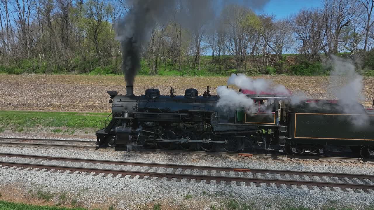 A historic steam locomotive travels down the tracks surrounded by vibrant spring greenery and blue skies. The engine releases clouds of steam, bringing nostalgia to railway enthusiasts