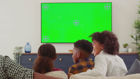 Rear-view-of-family-sitting-on-sofa-watching-green-screen-TV-at-home-as-father-jumps-in-surprise---shot-in-slow-motion