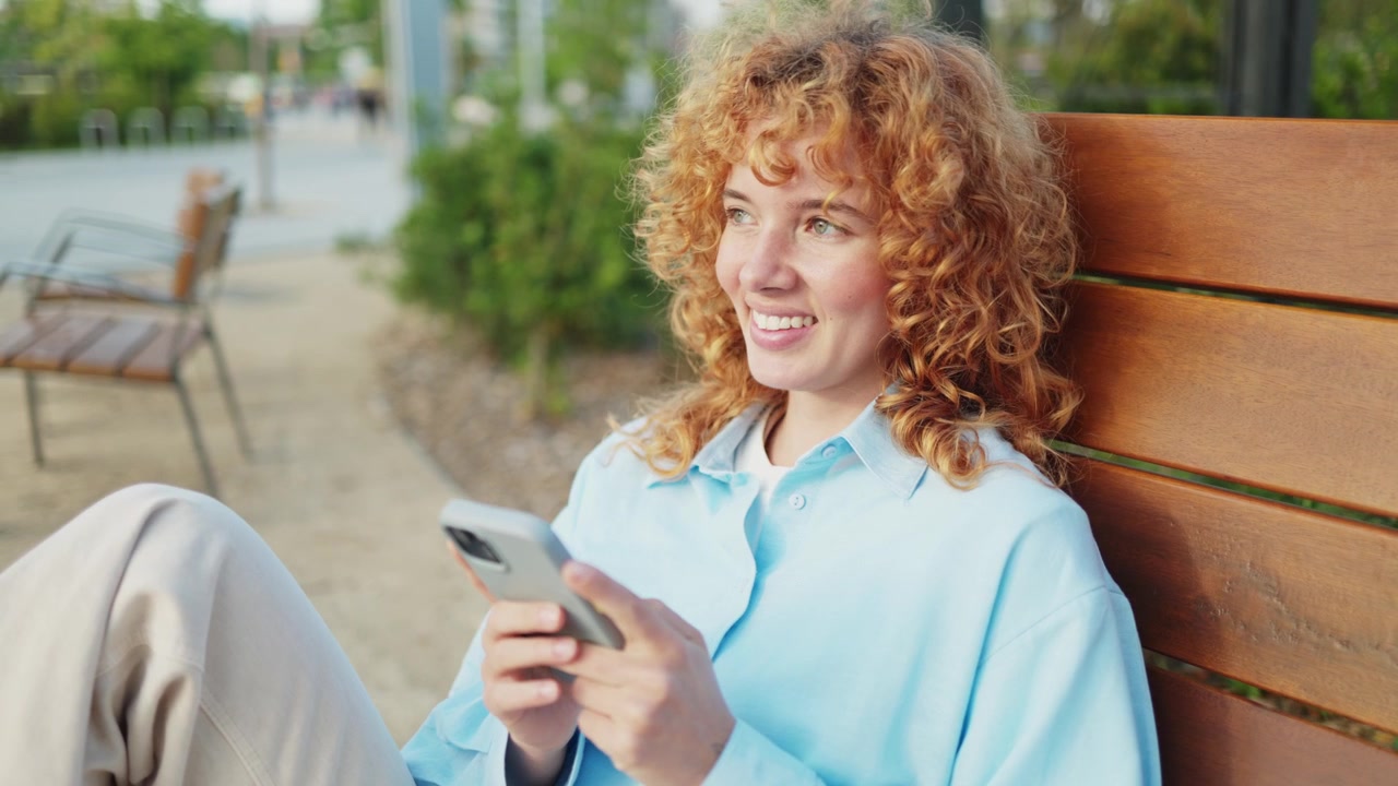 Smiling Young Woman with Curly Hair Using Smartphone on a Park Bench