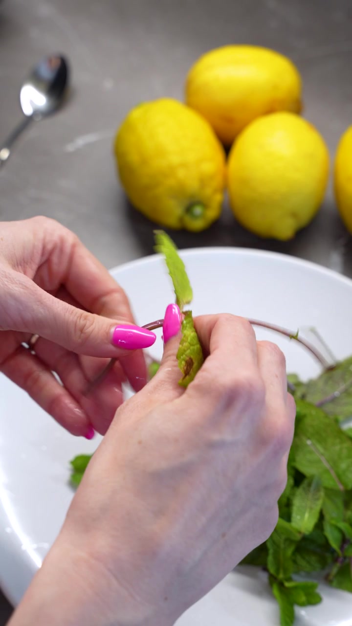 Primer plano vertical de manos femeninas deshojando hojas de menta fresca del tallo en un cuenco blanco. Al fondo se ven limones enteros para la preparación de una bebida saludable en una cocina.