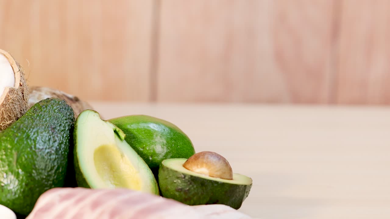 Various healthy fat sources, including avocado, coconut, olive oil, nuts, seeds, and butter, arranged on a wooden table in bright, natural lighting with a slow leftward camera pan