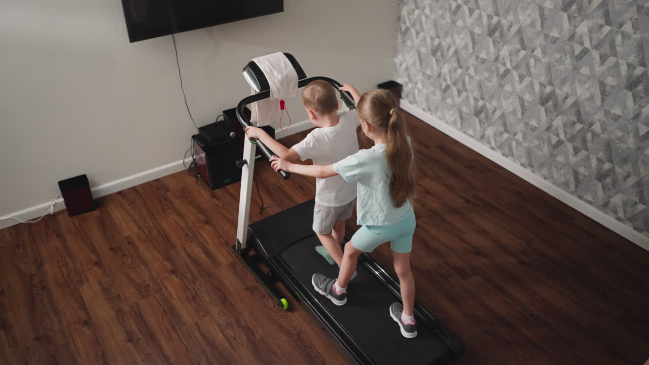 Rear view of young boy jumping slightly while holding treadmill handle during exercise as girl behind lets go of handle and tries to keep her balance in cozy room with wood flooring